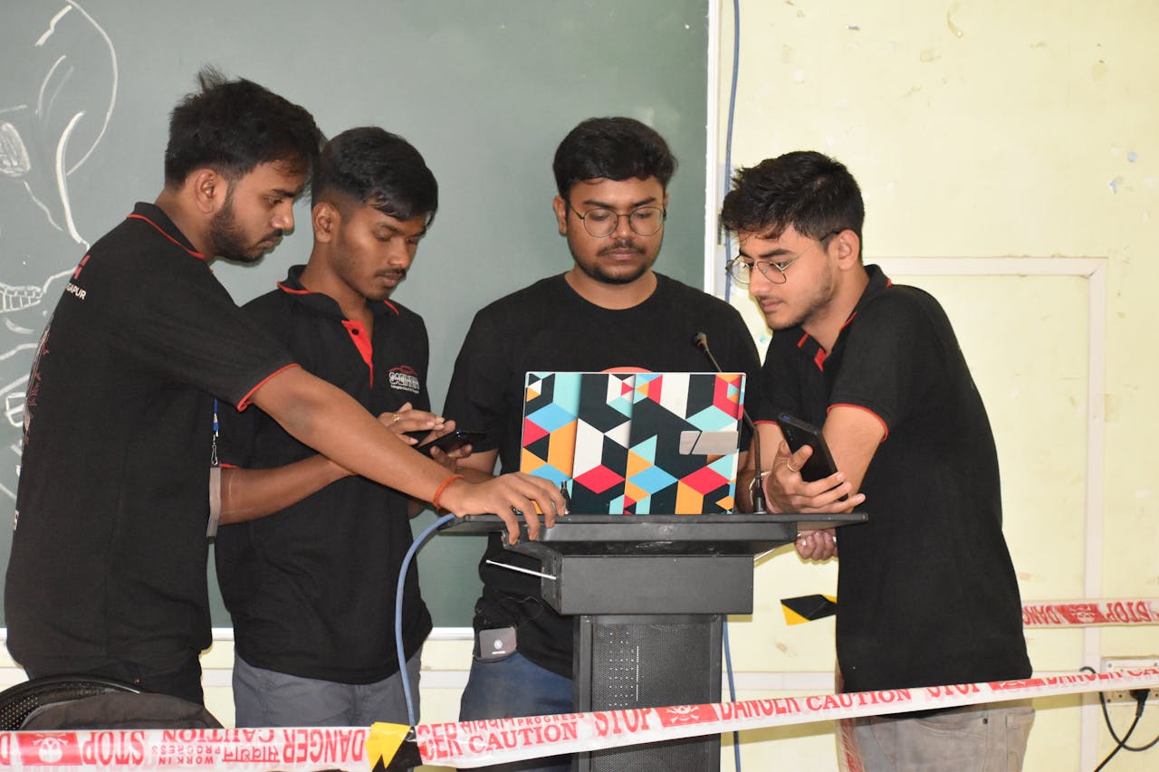 Four students work together on a laptop, preparing a presentation indoors by a chalkboard.